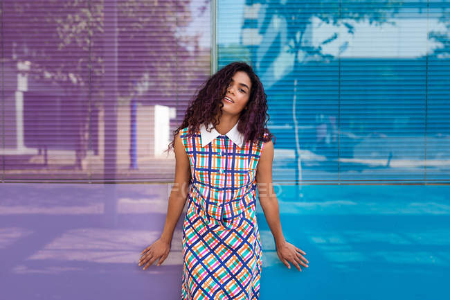 Sensual jovem etnia feminina em vestido colorido inclinado na parede de vidro de cor azul e rosa olhando para a câmera — Fotografia de Stock