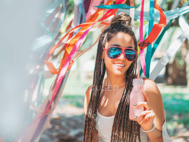 Elegante joven hembra con gafas de sol bebiendo cóctel durante el festival exterior y mirando a la cámara - foto de stock