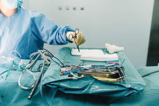 De arriba médico de cultivo en uniforme poner tijeras en bandeja con herramientas quirúrgicas inoxidables en quirófano - foto de stock