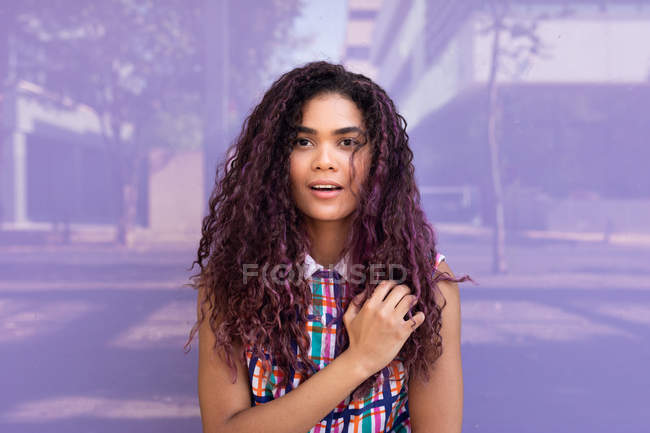 Portrait de charmante jeune femme ethnique aux cheveux bouclés regardant la caméra contre un mur de verre coloré — Photo de stock