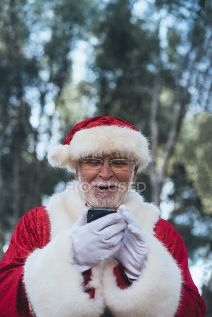 Homem alegre em traje de Papai Noel usando telefone celular moderno em fundo natureza turva — Fotografia de Stock