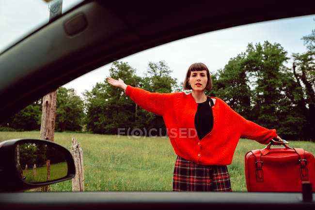 Femme en pull rouge gesticulant près de la voiture sur la route à la campagne — Photo de stock