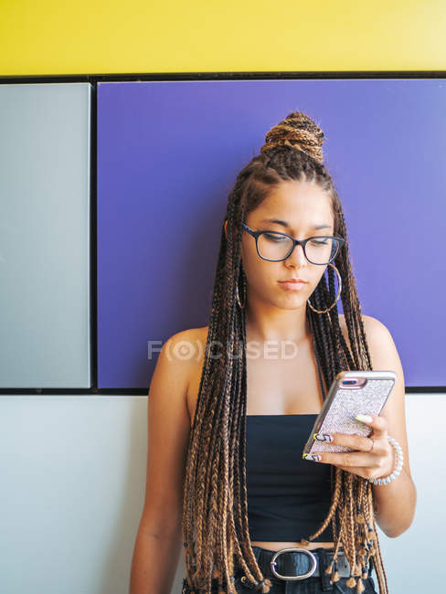 Jolie adolescente avec dreadlocks élégant en utilisant smartphone dans la chambre colorée — Photo de stock