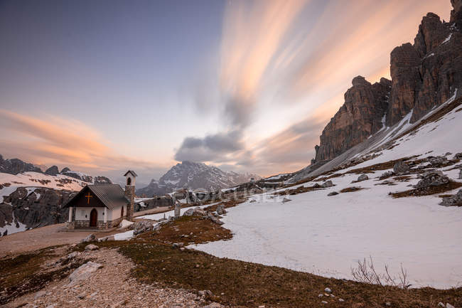 Petite église dans la vallée enneigée avec des montagnes au coucher du soleil — Photo de stock