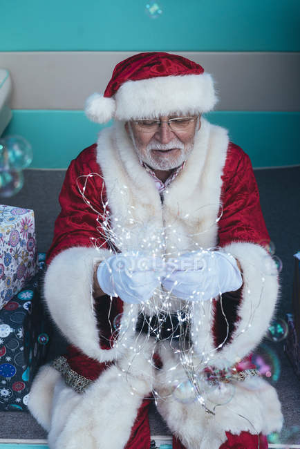 Senior man in costume of Santa Claus sitting in retro van and holding garland in gloved hands — Stock Photo