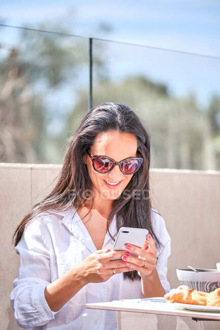Blogger sonriente sentada en la cafetería a la mesa con ensalada de verduras y tomando fotos de comida con smartphone - foto de stock