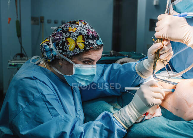 Vue latérale du jeune médecin sérieux en masque protecteur et capuchon faisant la chirurgie avec des instruments et infirmière de culture — Photo de stock