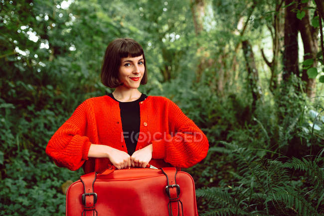 Woman in red with big vintage suitcase standing in forest and smiling — Stock Photo