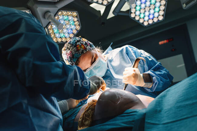 Médico joven serio en máscara protectora y cirugía de fabricación de gorras con instrumentos y enfermera de cultivos - foto de stock