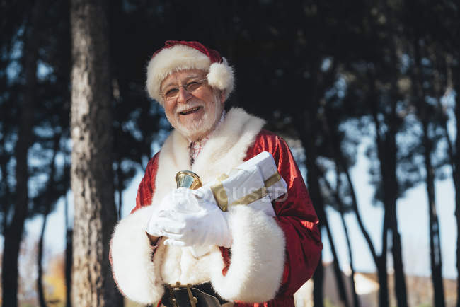 Веселий старший чоловік у костюмі Діда Мороза, стоячи з подарунком і дзвіночок в балакучих руках на фоні природи — стокове фото