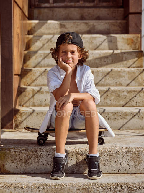 Niño pensativo sentado en el monopatín en las escaleras en skatepark en verano soleado mirando en la cámara - foto de stock