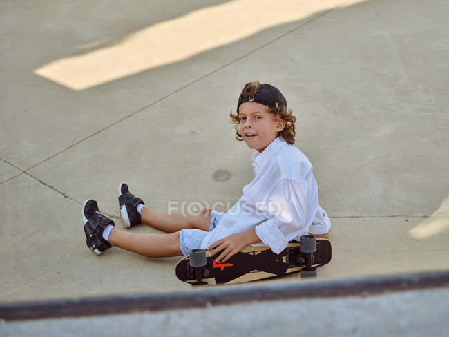 Criança feliz sentado e arrepiante no chão com skate no parque de skate olhando para a câmera — Fotografia de Stock