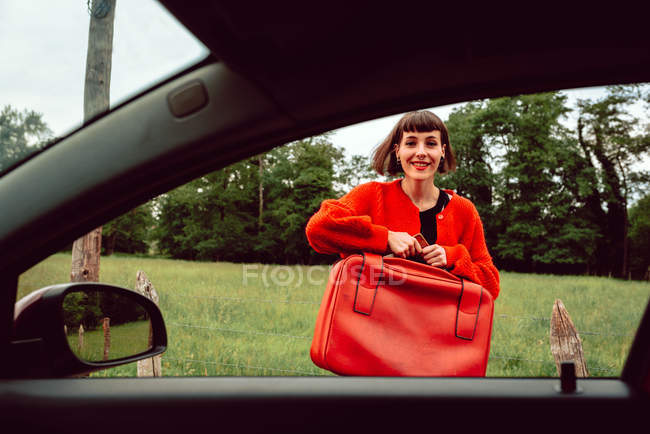Усміхнена молода жінка кладе валізу перед вікном автомобіля — стокове фото