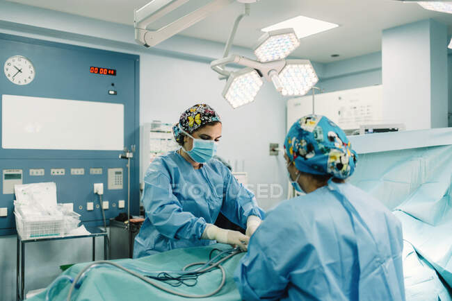Medics in blue surgical gowns and protective masks holing pipes in body of covered patient lying on bed — Stock Photo