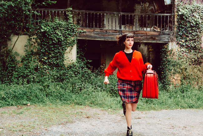 Mujer joven en rojo con gran maleta roja corriendo en el campo contra casa abandonada - foto de stock