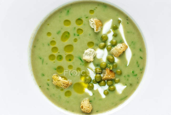 De arriba la sopa de cremas vegetales enmascarada con guisantes verdes y pequeños crackers sobre fondo blanco. - foto de stock