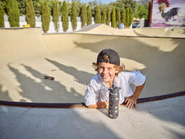 Cool petit garçon dans le capuchon accroché sur la rampe de patinage et l'eau potable de la bouteille de sport avec de la paille regardant à la caméra — Photo de stock