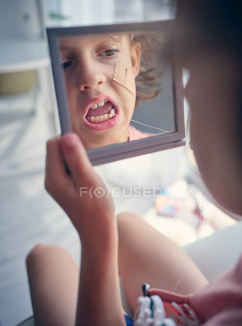 Відбиття в квадратному дзеркалі обличчя дитини з бинтом на щоці вивчає молочний зуб з відкритим ротом в кімнаті — стокове фото