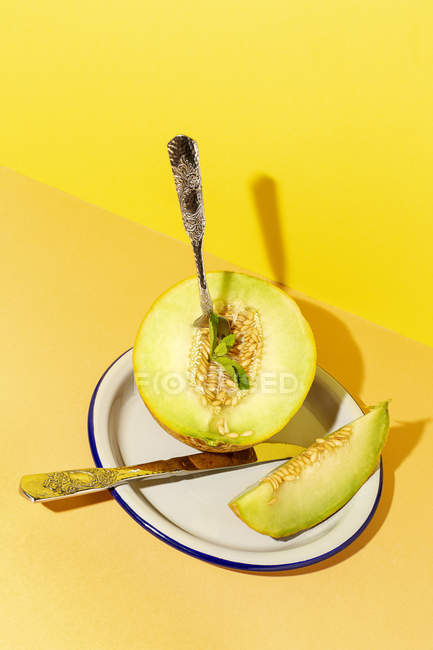 Cut ripe appetizing sweet pitted melon on plate with spoon and fork on yellow and orange background — Stock Photo