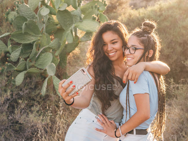 Deliziose donne felici casual scattare foto su smartphone mentre in piedi accanto a cactus in campagna deserta al tramonto — Foto stock