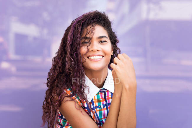 Ritratto di giovane donna etnica sorridente con i capelli ricci che guarda la macchina fotografica contro la parete di vetro viola — Foto stock