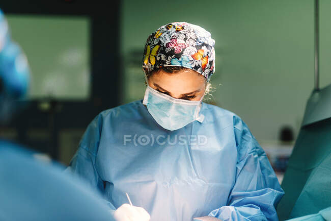 Jovem médico grave com máscara protetora e tampa fazendo cirurgia com instrumentos e enfermeira de colheita — Fotografia de Stock