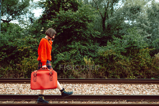 Stylish fearless woman with bright red suitcase stepping on railroad ties in countryside — Stock Photo