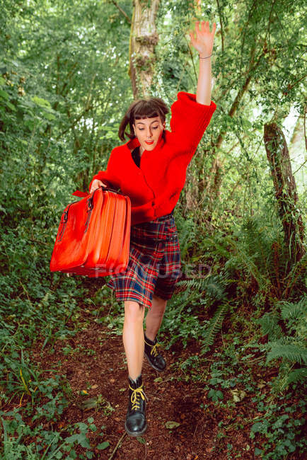 Жінка в червоному з великою старовинною валіза біжить в зеленому лісі — стокове фото