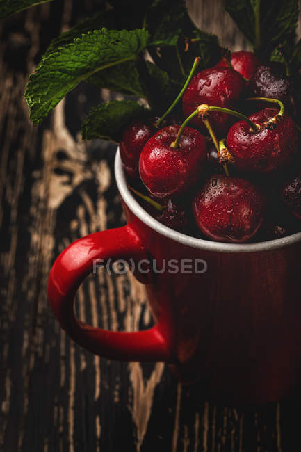 Sabrosas y apetitosas cerezas maduras con hojas en taza roja sobre una mesa de madera oscura - foto de stock