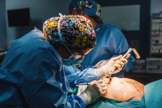 Сторона зору лікаря в хірургічній сукні і захисній масці, що зрізає груди пацієнта лежить на ліжку. — Stock Photo