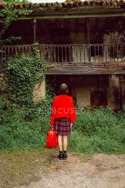 Woman in red with big vintage red suitcase standing in front of abandoned house in countryside — Stock Photo
