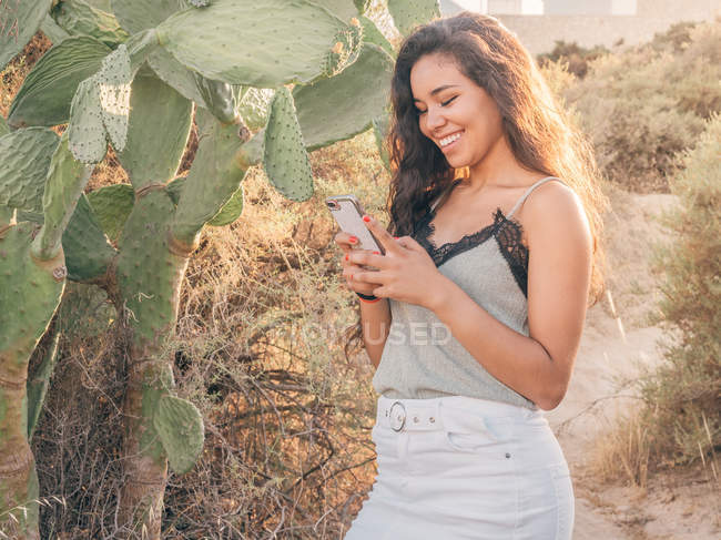 Femme naviguant sur smartphone tout en se tenant à côté de cactus sur le chemin sablonneux — Photo de stock