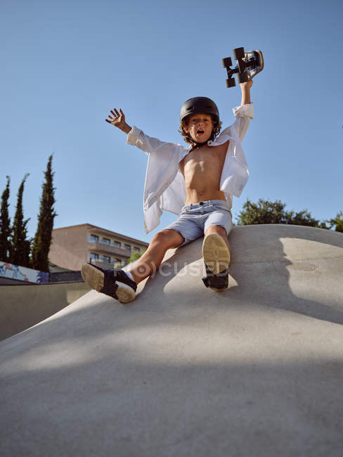 Dal basso del ragazzo felice con il casco che tiene lo skateboard sopra la testa e urla mentre siede sulla rampa contro il cielo blu — Foto stock