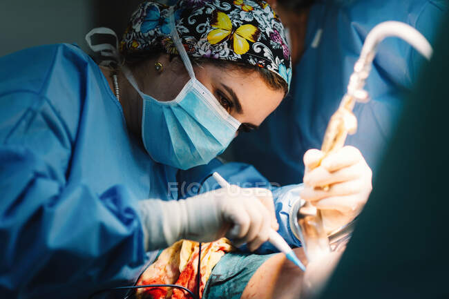 Vista lateral del médico joven serio en la máscara protectora y la toma de la tapa de la cirugía con instrumentos y enfermera de cosecha - foto de stock