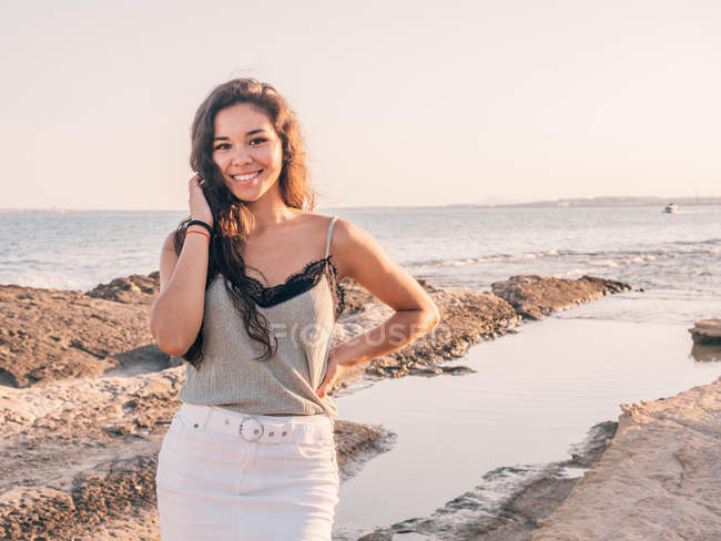 Contenuto sorridente donna casual in piedi in spiaggia accanto alla costa alla luce del sole — Foto stock