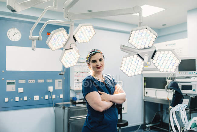 Доросла жінка в медичній формі перетинає руки і дивиться на камеру, стоячи біля ламп в сучасному операційному театрі — стокове фото