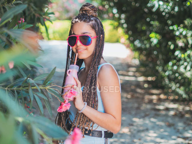 Усміхнена молода жінка в сонцезахисних окулярах з дредлоками п'є коктейль і дивиться на камеру в парку — стокове фото