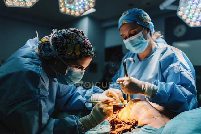 Seitenansicht eines ernsthaften jungen Arztes in Schutzmaske und Mütze, der mit Instrumenten und einer Krankenschwester operiert — Stockfoto