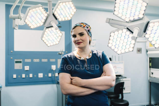 Доросла жінка в медичній формі перетинає руки і дивиться на камеру, стоячи біля ламп в сучасному операційному театрі — стокове фото
