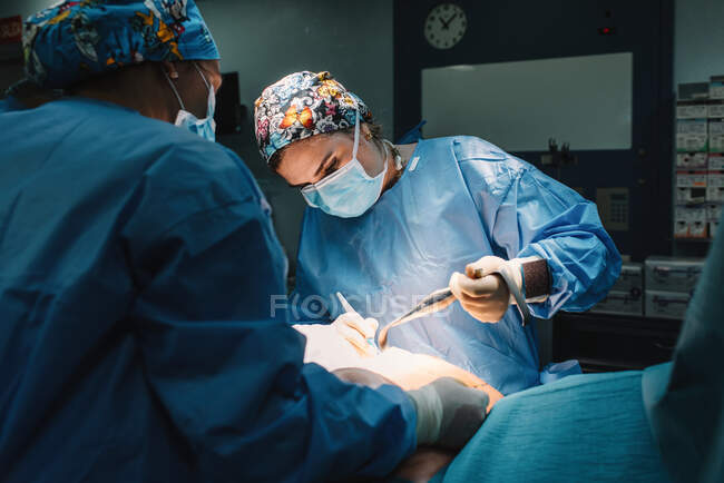 Sérieux jeune médecin en masque protecteur et casquette faisant la chirurgie avec des instruments et infirmière de culture — Photo de stock