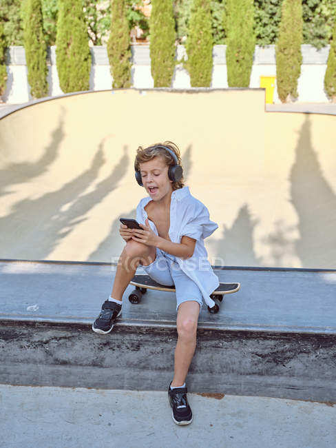 Мальчик в наушниках и с помощью смартфона сидит на скейтборде в солнечном городском скейтпарке — стоковое фото
