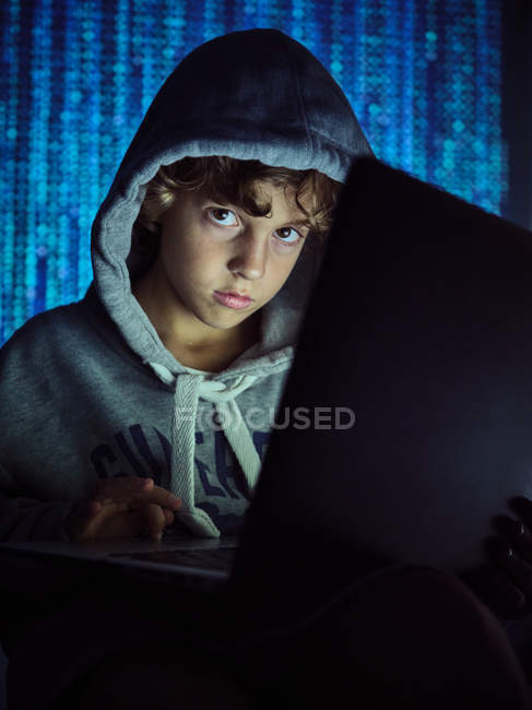 Niño en sudadera con capucha mirando a la cámara y utilizando el ordenador portátil para robar información en el ciberespacio - foto de stock