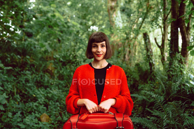 Mulher em vermelho com grande mala vermelha andando na floresta verde — Fotografia de Stock