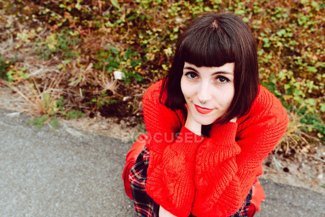 Молодая стильная женщина сидит на красном чемодане на дороге и смотрит в камеру — стоковое фото