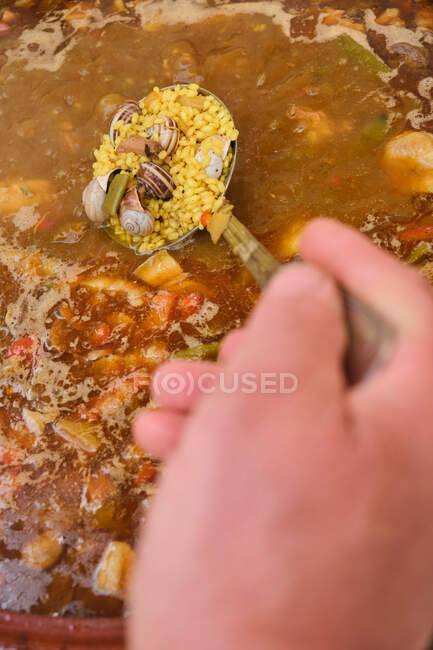 Cultivo anónimo de pie humano poner cuchara en una olla de barro grande con plato de cocina de arroz de pollo surtido de verduras y caracoles - foto de stock