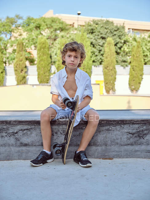 Sorrindo garoto casual em capacete e camisa branca sentado na rampa no parque de skate olhando na câmera — Fotografia de Stock