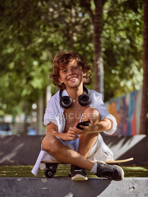 Случайный задумчивый мальчик в наушниках с помощью мобильного телефона сидит на скейтборде во время отдыха в скейтпарке — стоковое фото