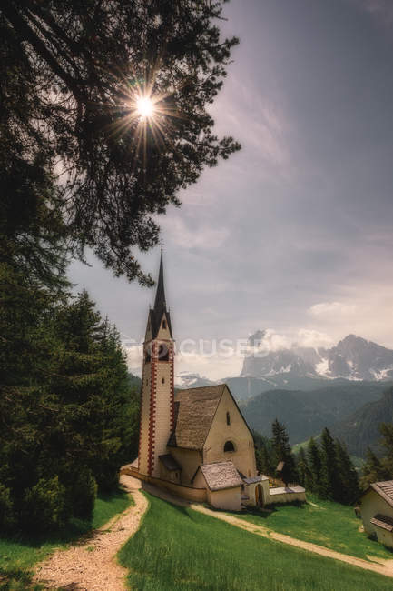 Petite église sur une colline verdoyante sous un soleil éclatant et des montagnes rocheuses en arrière-plan à Dolomites, Italie — Photo de stock