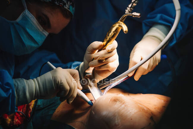 Visão lateral do jovem médico grave em máscara protetora e tampa fazendo cirurgia com instrumentos e enfermeira de colheita — Fotografia de Stock