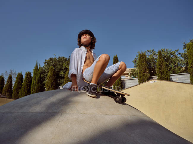 Снизу счастливый мальчик в шлеме держа скейтборд сидя на рампе против голубого неба — стоковое фото
